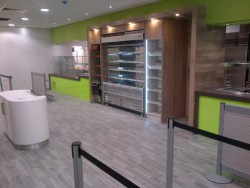 Bespoke counters with silestone worktops including drop to floor sections, frontage finished in green acrylic, central retail section finished in wood effect laminate with LED strip lighting on retail housing, grey wood effect vinyl flooring